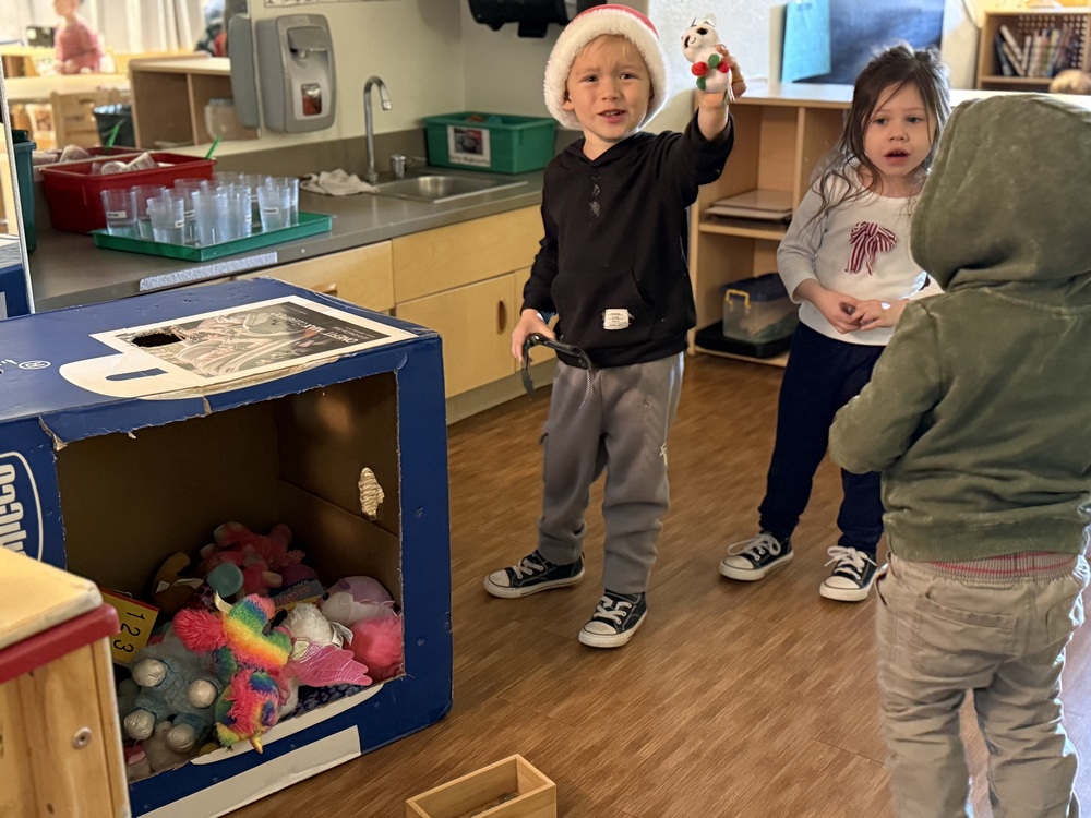 Three students in their classroom stand next to a cardboard box they have made into a pretend arcade claw machine with stuffed animals inside. 