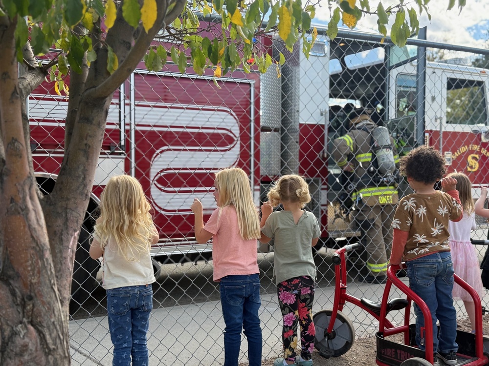 Students look at the firemen and fire truck through the fence on the playground at the Early Childhood Center.
