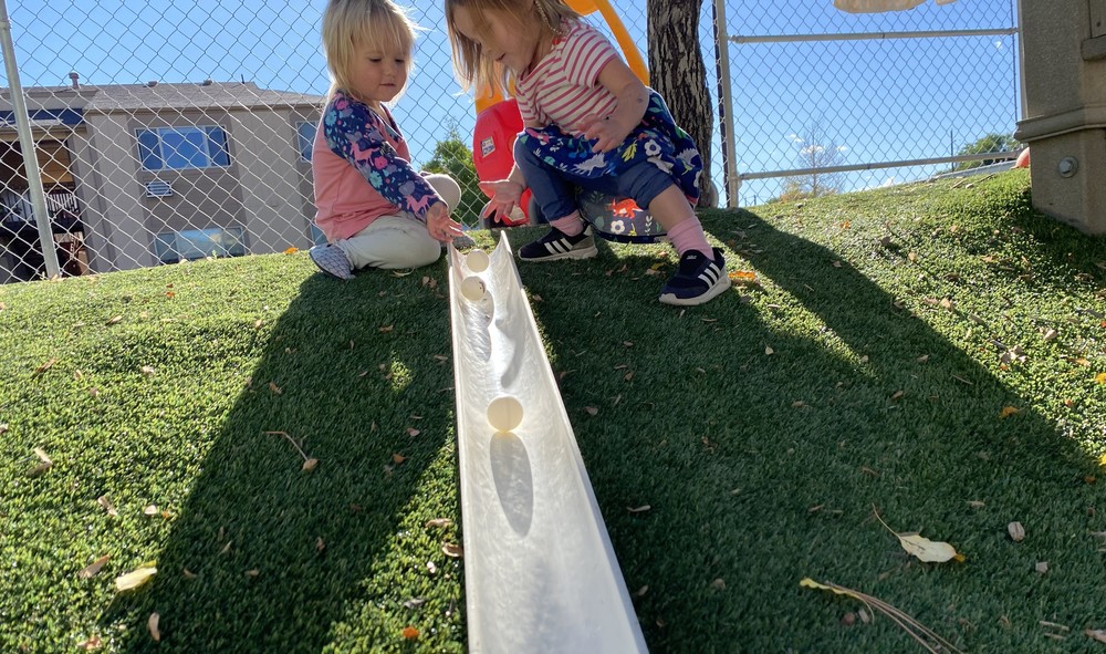 Two students roll balls down a ramp on the toddler plaground