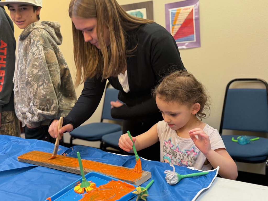 A middle school student and a preschool student paint a piece of wood orange.