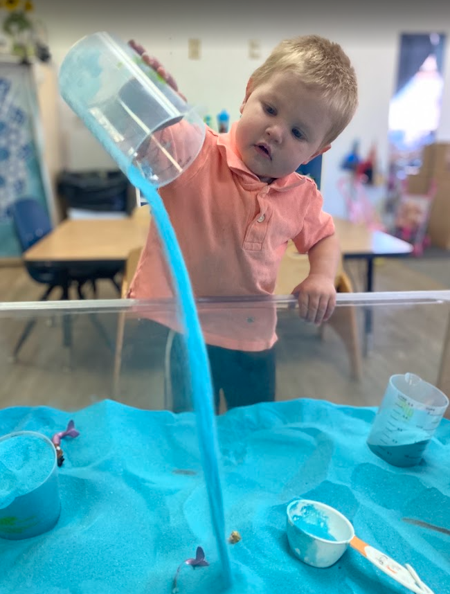 A ECC student pours blue sand out of a pitcher into the sensory table in his classroom. 