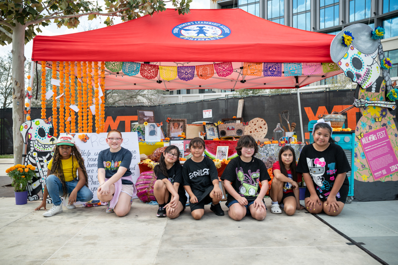 Girls sit in front of large altar with catrina at stove