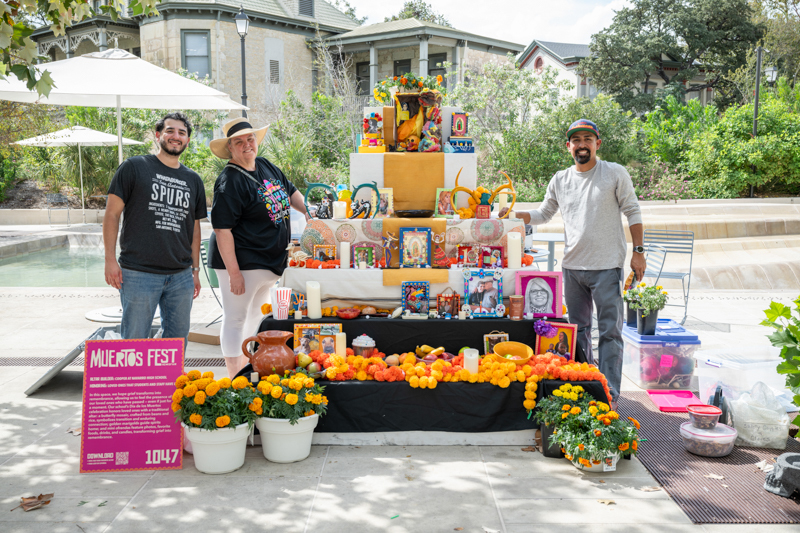 Three adults stand next to multi-tiered altar with marigolds