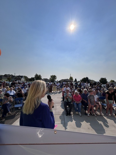 Bright sun in a clear sky. A woman speaks into a microphone while a crowd of people sits and stands.