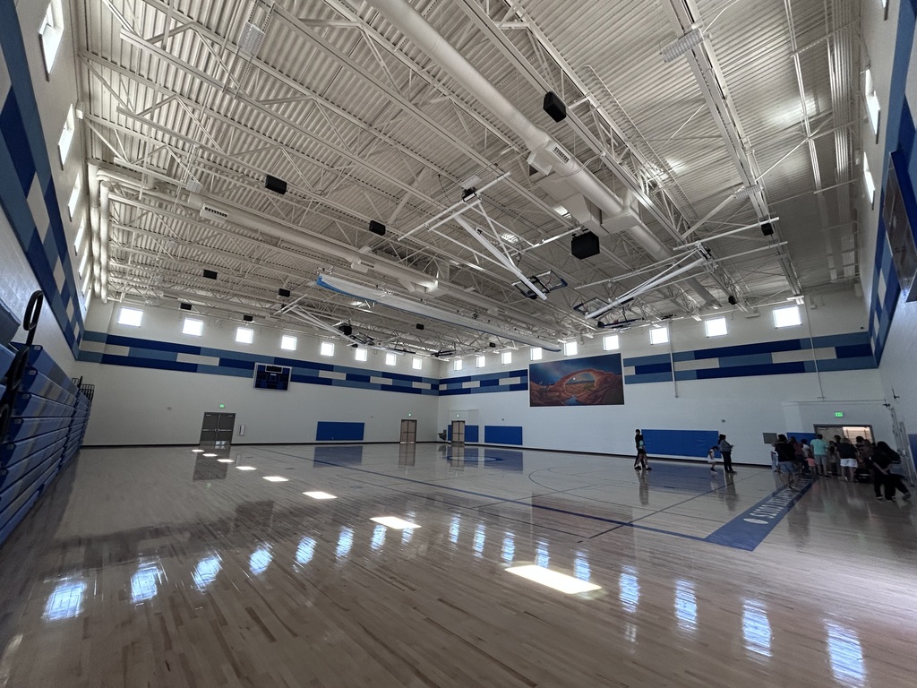 An indoor basketball court with empty bleachers, white and blue walls, and a white ceiling. A person is sitting on a bench.
