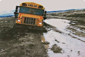 A yellow school bus rides through a muddy puddle. The bus has a driver behind the wheel.
