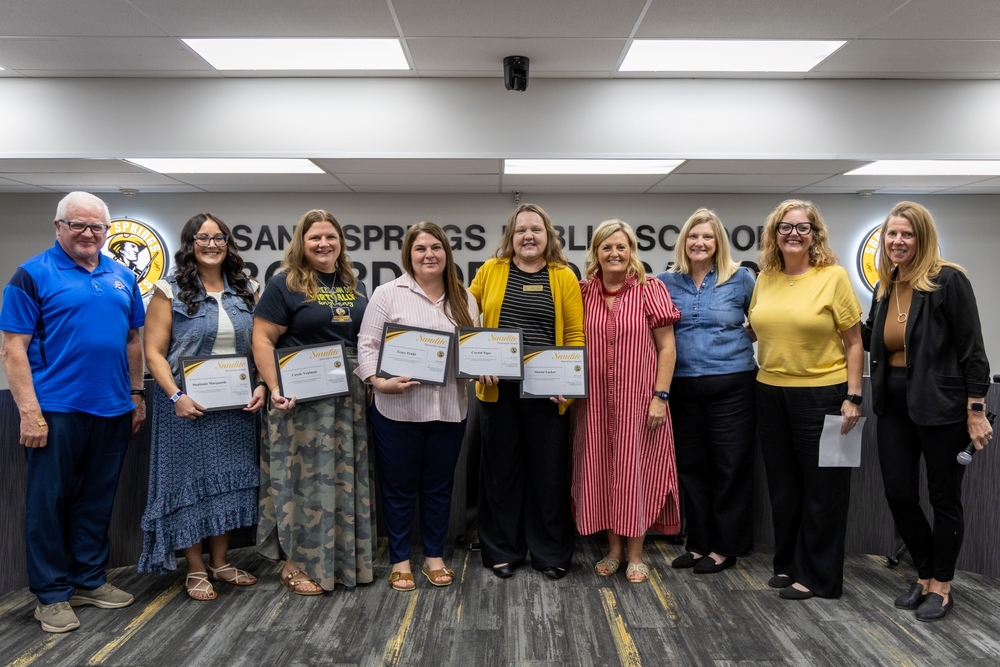 A group of four honorees and five school officials posing with framed Sandite Pacesetter Award certificates in front of the Sand Springs Public Schools Board of Education wall.