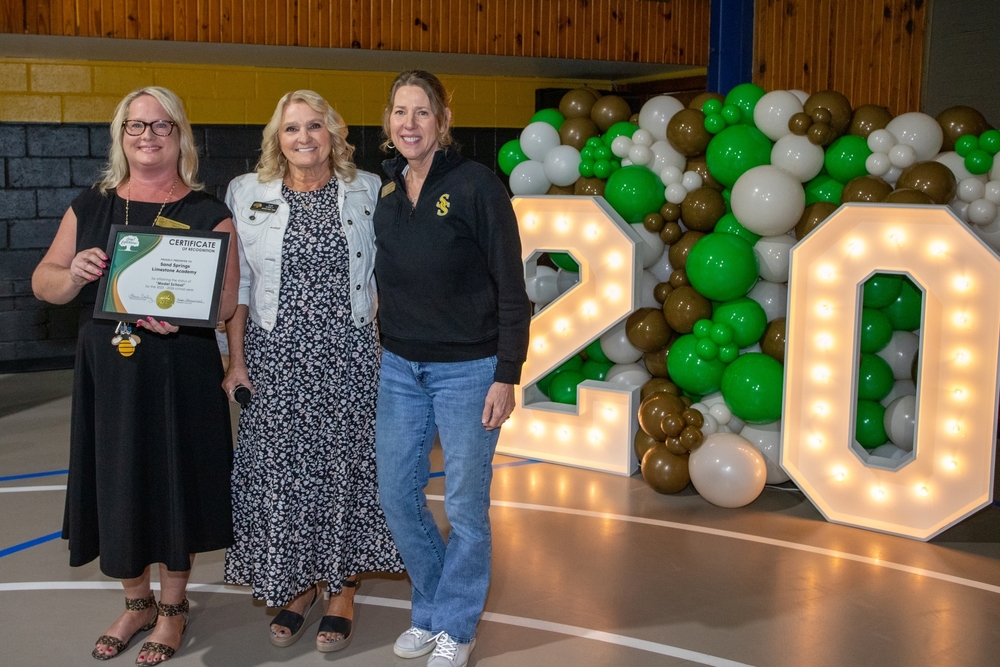 Three women stand together indoors during an award presentation while one holds a framed certificate of recognition. Behind them is a large green white and gold balloon display with illuminated numbers reading 20, suggesting a twentieth anniversary or milestone celebration.