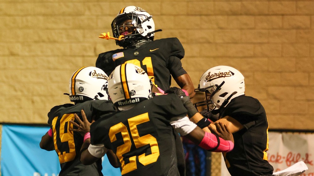 A group of Sand Springs football players in black uniforms and white helmets lift a teammate into the air in celebration during a game.