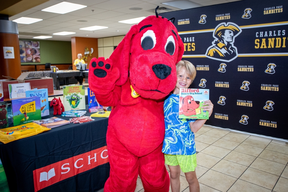 Student smiles while posing beside the Clifford the Big Red Dog mascot and holding a copy of “Clifford Goes to Dog School.” A display table filled with children’s books and a Charles Page Sandites backdrop frame the scene during a school literacy event celebrating reading and books.