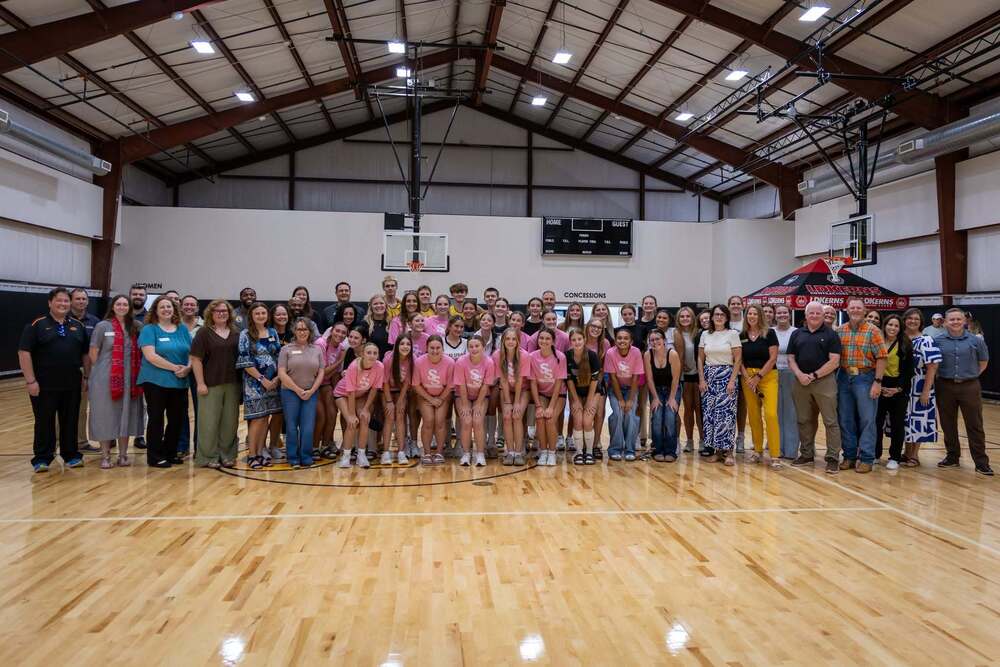 A large group of people—including students, staff, and athletes in matching pink shirts—pose together on a polished indoor basketball court.