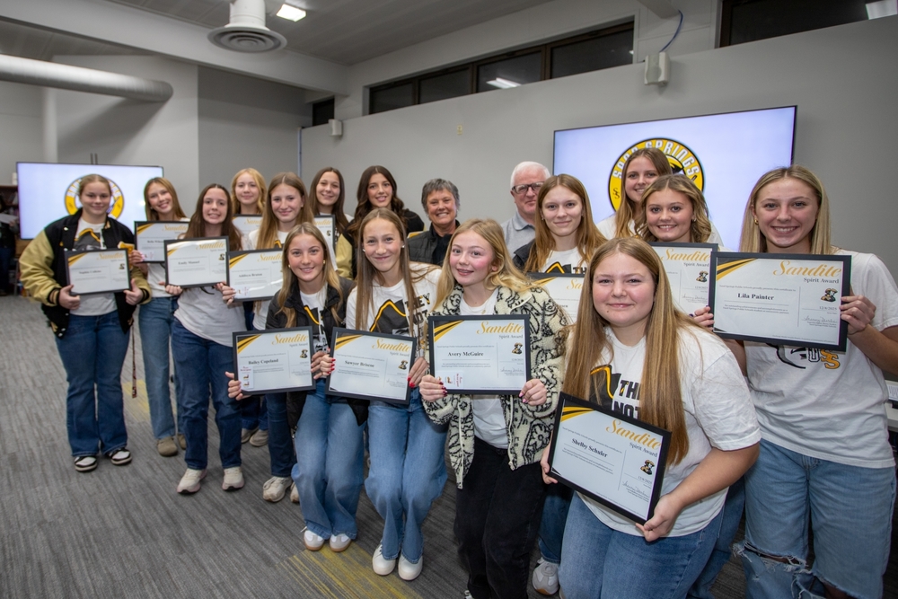 A large group of Lady Sandites athletes stand together at a Board of Education meeting, each holding a Sandite Spirit Award certificate. Coaches and board members stand behind them, and Sand Springs Sandites logos are displayed on screen in the background.