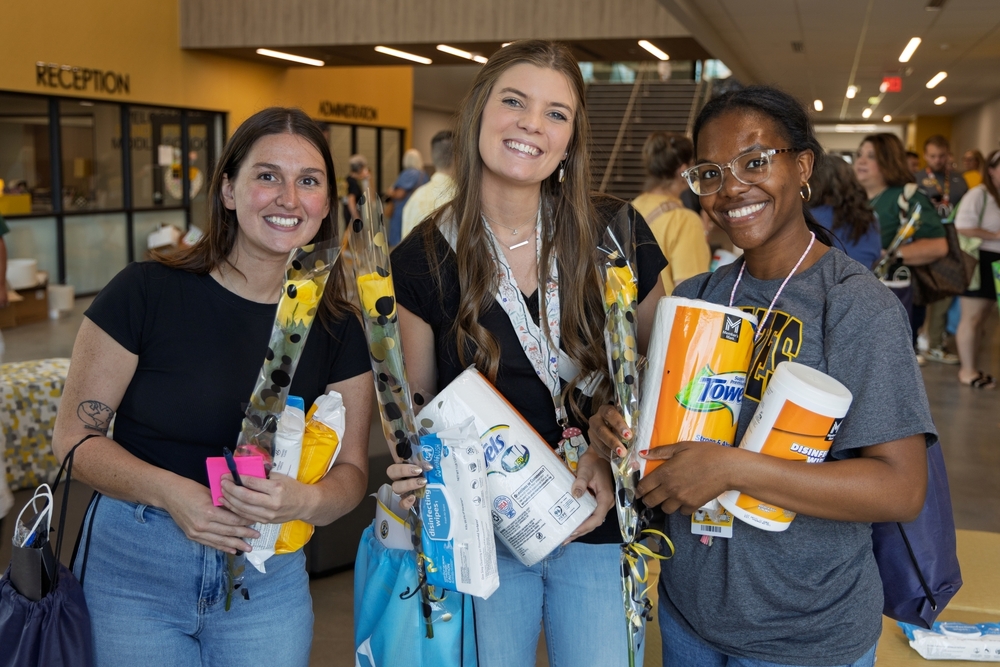 Three smiling women stand together indoors holding yellow roses, paper towels, and containers of disinfecting wipes.