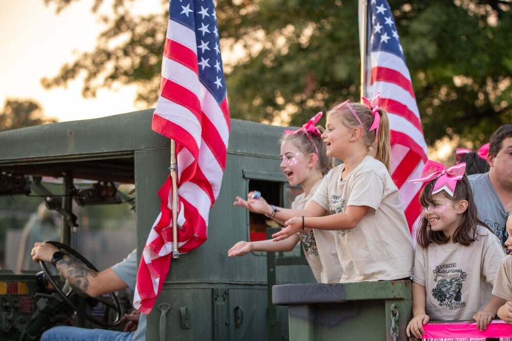 Three young girls wearing matching beige Sandite Cheerleading shirts with pink hair bows lean out from the back of a green military truck during a parade, tossing candy while American flags flutter beside them.