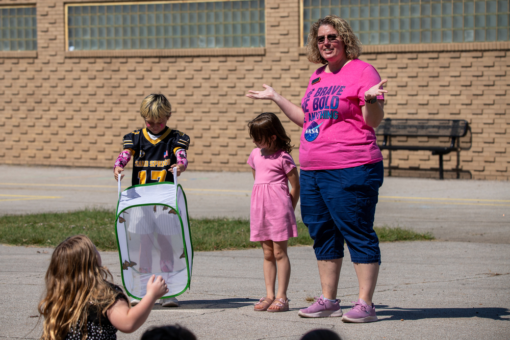 A teacher stands outside with two students during a sunny outdoor activity. The teacher, wearing a bright pink T-shirt and blue shorts, gestures with open hands while smiling. One student in a Sand Springs football jersey holds a mesh enclosure containing butterflies, and another student in a pink dress stands beside them. Other children sit on the pavement watching the demonstration in front of a brick school building.