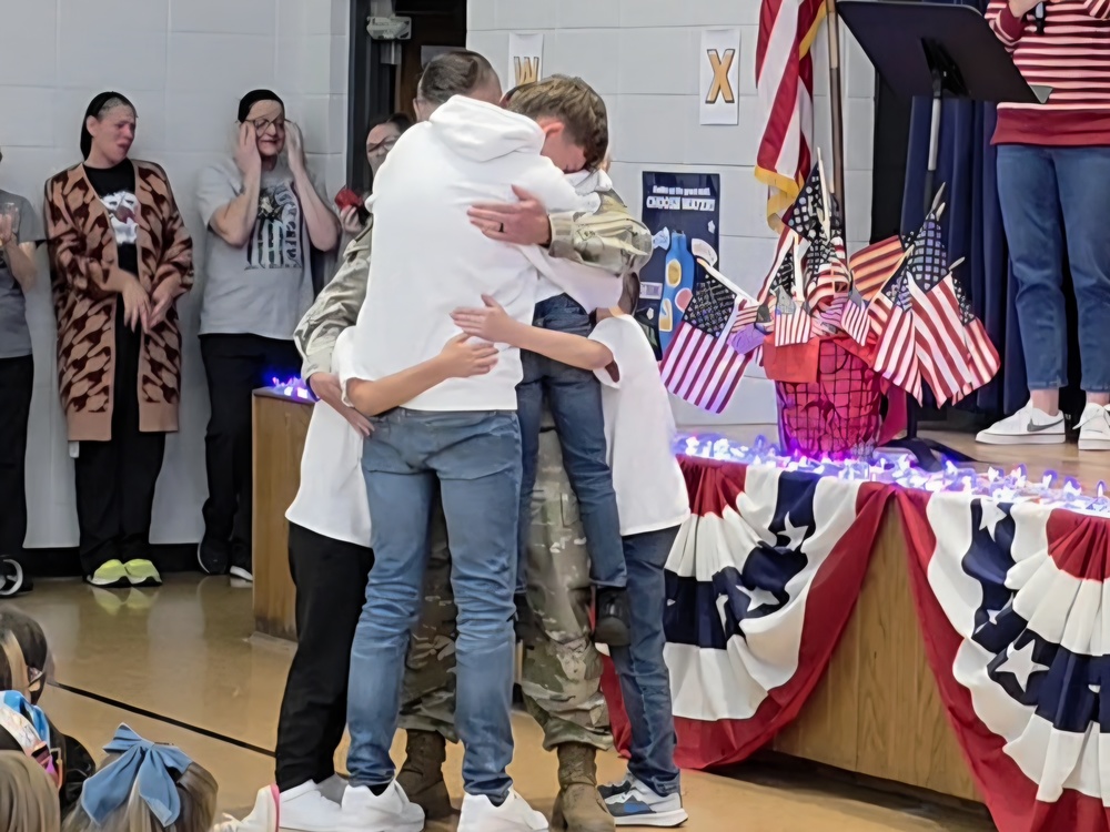 A soldier in uniform embraces three children and a teenager in an emotional reunion during a school assembly. The stage behind them is decorated with patriotic red, white, and blue bunting and American flags, while teachers and staff look on with tears and smiles.