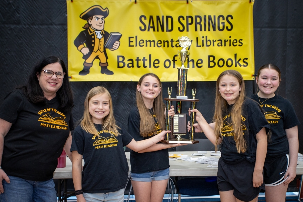 Group of elementary students holding a trophy. Banner in the background that reads "Sand Springs Elementary Libraries Battle of the Books".