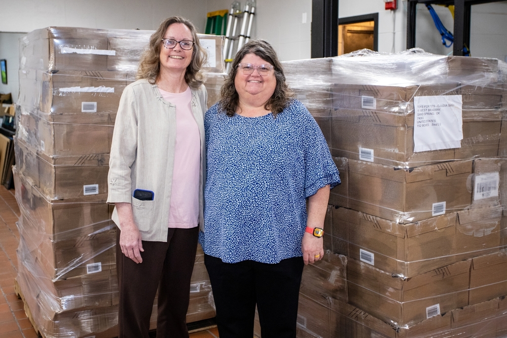 Two school administrators standing next to shrink-wrapped pallets of boxes.