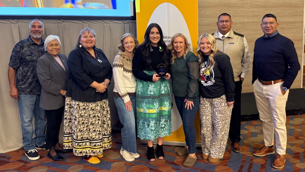 A group of nine people stand together smiling at an award event. The woman in the center holds a glass plaque recognizing an achievement. The group stands in front of a yellow and white banner with the Johnson O’Malley Program logo visible in the background.