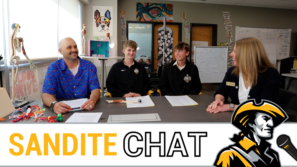 High school students and school officials sit at a table in the biomedical classroom. DNA models and anatomical displays decorate the room. The words "Sandite Chat" next to the Sandite minuteman logo and a microphone.