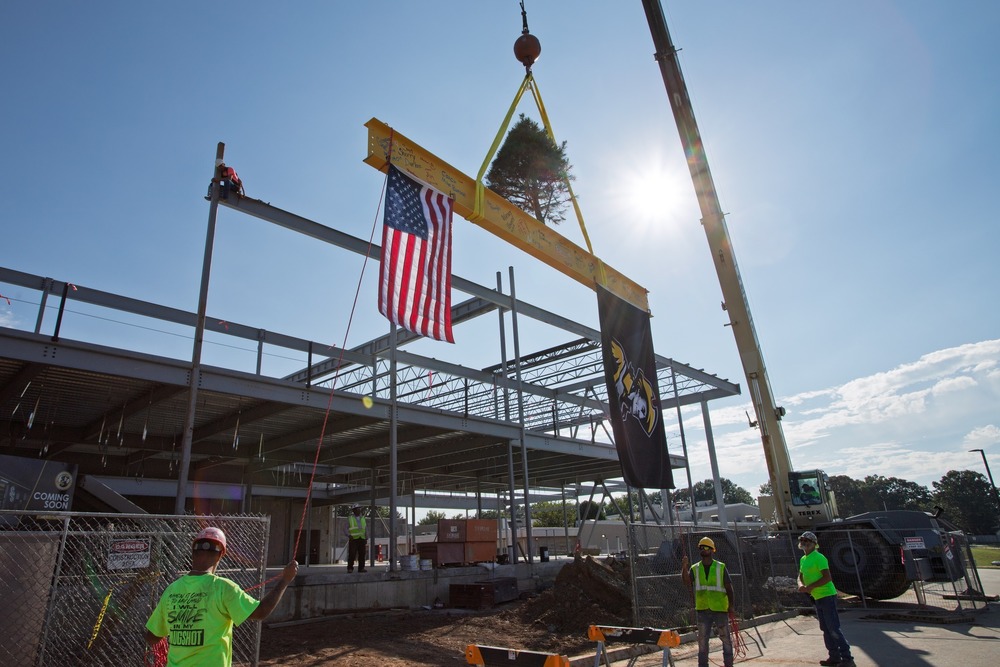 A crane lifts the signed yellow beam with flags and a tree attached above a building under construction.