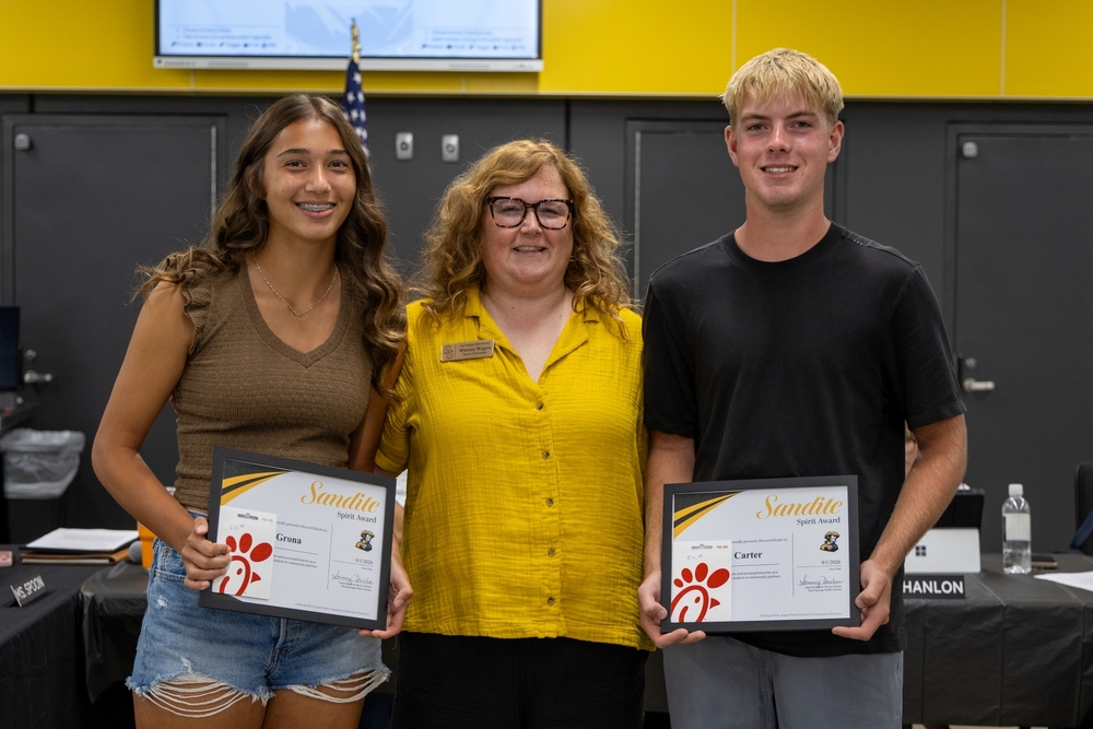 An adult woman in a yellow shirt stands between two high school students holding Framed Sandite Spirit Award certificates and Chick-fil-A gift cards.