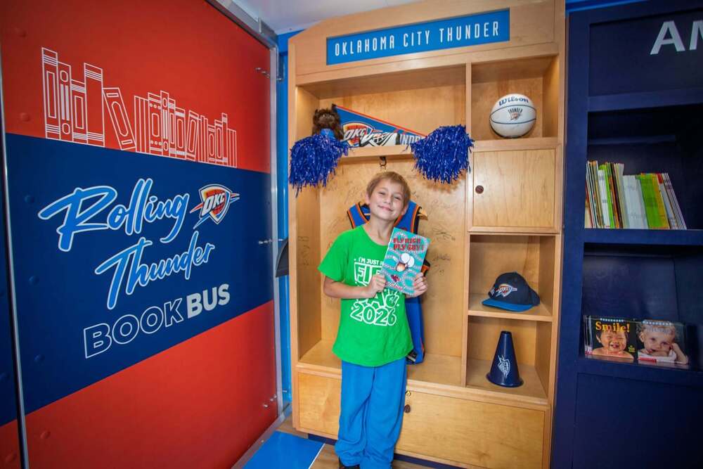 Student wearing a bright green “I’m Just Here for Field Day 2026” shirt stands inside the Oklahoma City Thunder Book Bus holding a copy of “Fly Guy! Fly Guy!” Behind the student is a Thunder-themed reading nook decorated with team memorabilia, including pom-poms, a basketball, a pennant, and a sign reading “Oklahoma City Thunder,” while a wall graphic reads “Rolling Thunder Book Bus.”