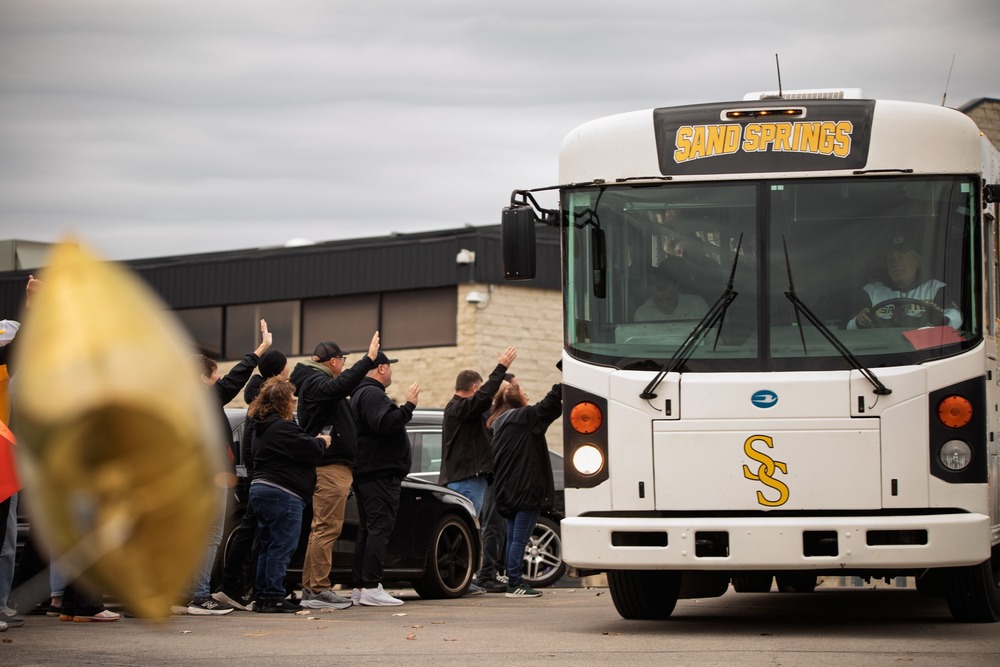 Community members line the curb and wave as a Sand Springs team bus passes by outside a school building.