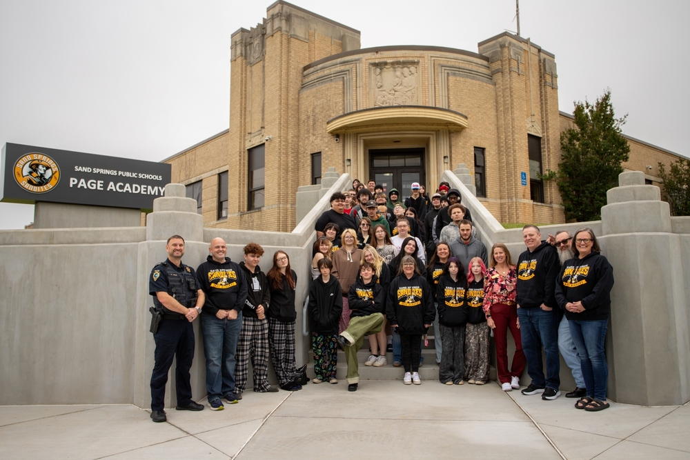 Large group of students, staff, and community members stand on the steps outside Sand Springs Public Schools Page Academy for a group photo. A sign with the Sandites logo reads “Sand Springs Public Schools Page Academy” beside the historic brick school building as participants smile toward the camera.