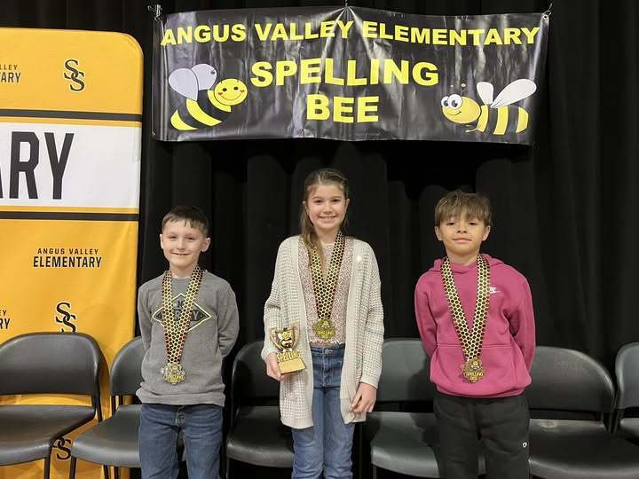 The top three spellers stand in front of the Spelling Bee banner on the stage. Each student wears a hedal and the winner, in the center, holds her Spelling Bee trophy.