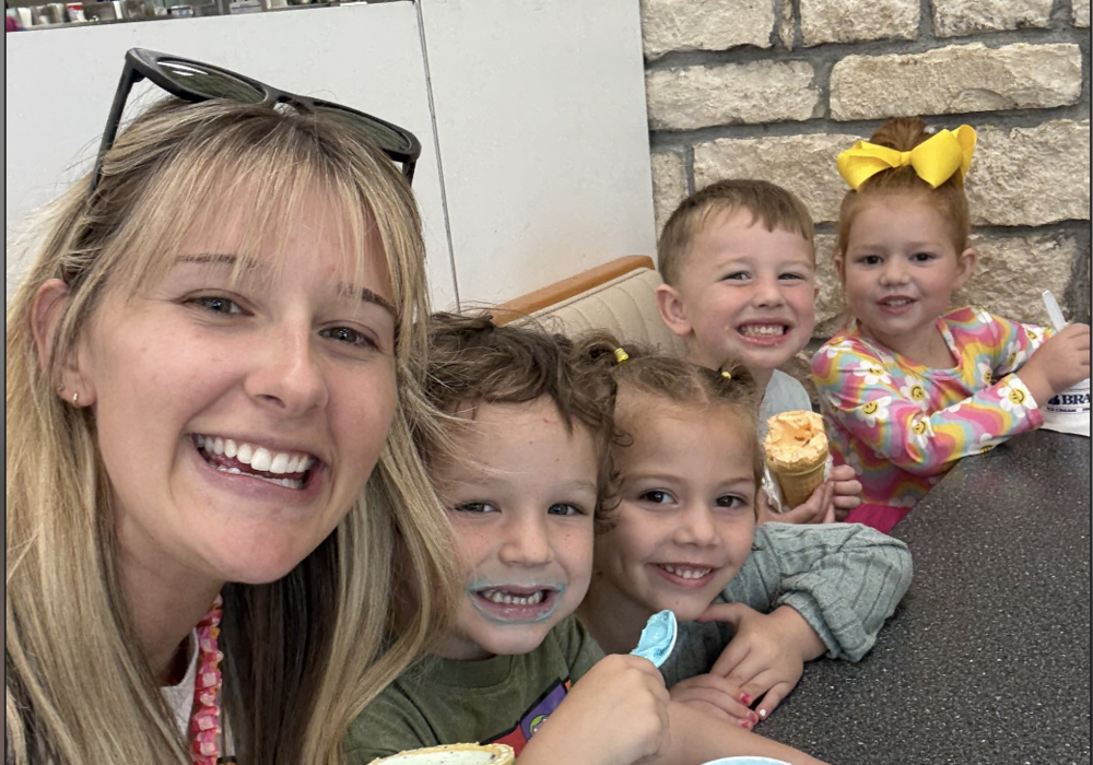 A smiling teacher and four young children sit together at a table inside Braum's, happily enjoying ice cream cones and cups. The children are laughing and holding colorful ice cream, with a stone wall in the back.