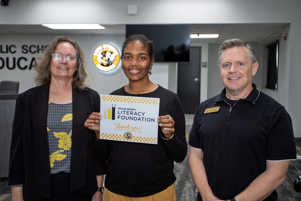 Claudia Smith, Robin Harris, and Shawn Beard pose for a photo. Harris holds a sign with the Dollar General Literacy Foundation logo and the words "Thank you!" above the Sand Springs Sandites seal.