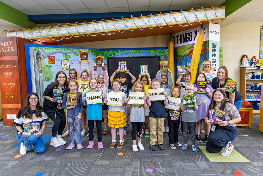A group of elementary school students and teachers smile and pose in a library while holding up books and signs that read "THANK YOU DOLLAR GENERAL."