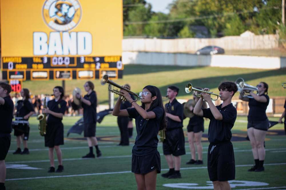High school marching band members in black uniforms perform on a football field, with two trumpet players in the foreground and a large yellow scoreboard reading "BAND" in the background.