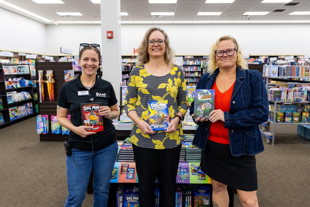 Three smiling women stand in a bookstore holding children's books, including two titles from the Dog Man series.