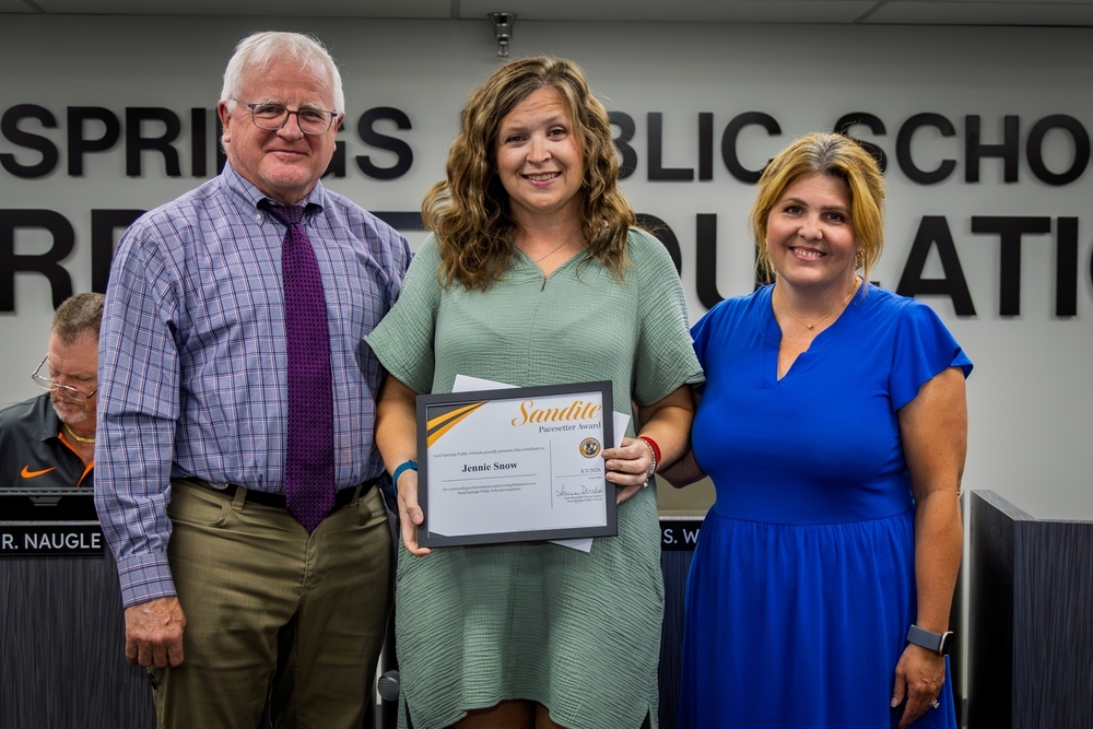Jennie Snow receives the Sandite Pacesetter Award framed certificate alongside a board member and administrator at a Sand Springs Public Schools board meeting.