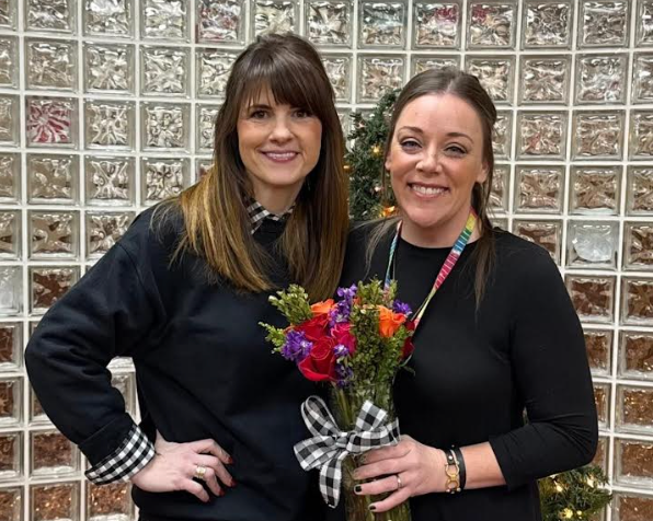 Principal Lisa Banks stands beside ECEC Teacher of the Year Jonelle Jones, who is holding a colorful bouquet of flowers tied with a black-and-white bow. Both women are smiling and dressed in black, posing indoors in front of a glass block wall decorated with holiday greenery and lights.