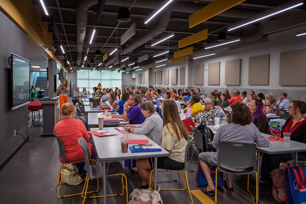 A large group of educators sits at tables in a conference-style training room while a presenter speaks at the front beside a large display screen. Participants listen attentively with notebooks, folders, and learning materials spread across the tables during a professional development session.