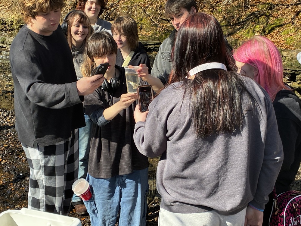 Students outdoors examine a trout in a clear container.