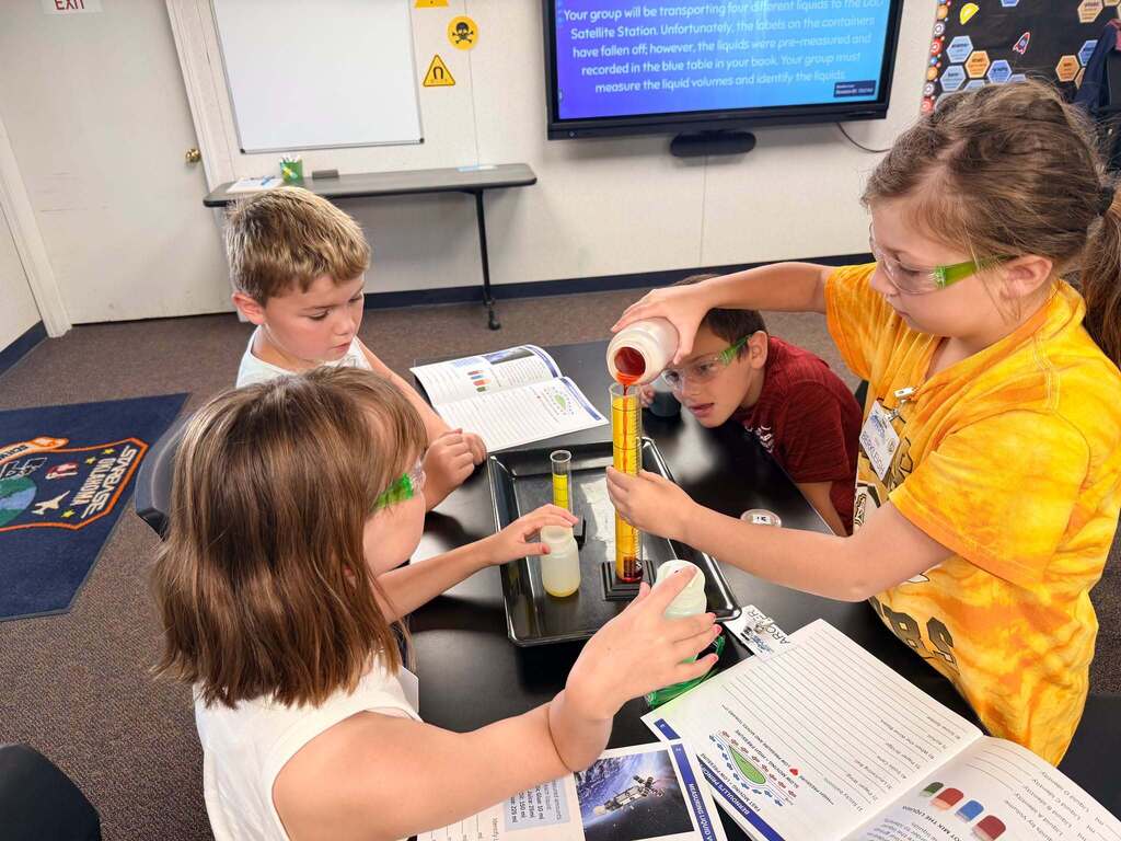 Four students wearing safety glasses gathered around a table as one pours red liquid from a container into a graduated cylinder.