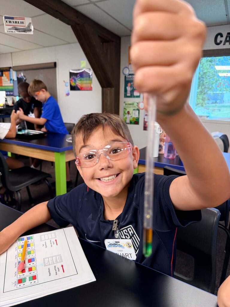A smiling student wearing safety glasses holds up a clear pipette filled with layered colored liquids.