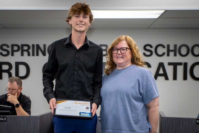 Student stands beside a board member while holding a framed “Sandite Spirit Award” certificate during a Sand Springs Public Schools Board of Education recognition ceremony. Both smile toward the camera in the board meeting room, with board members seated in the background and large wall lettering visible behind them.