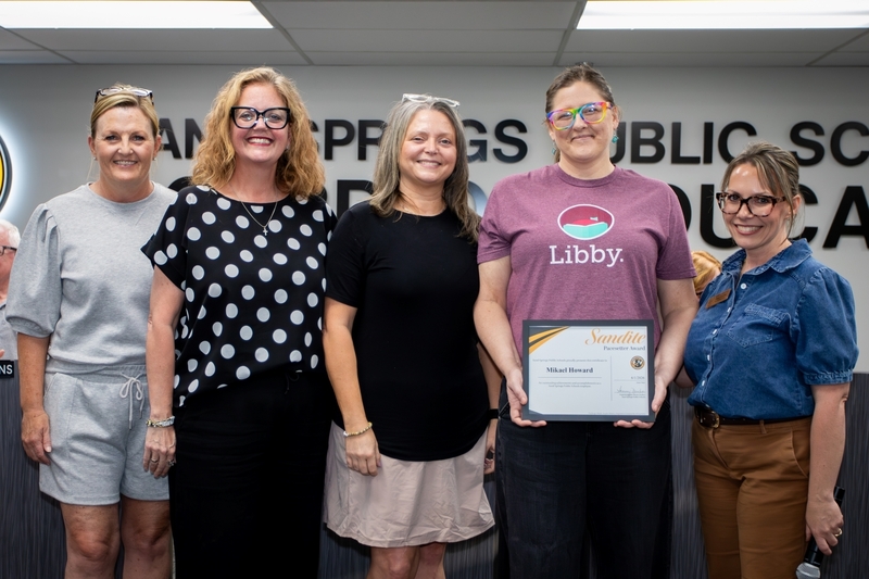 Four staff members and one board member stand together during a Sand Springs Public Schools Board of Education recognition ceremony. One recipient holds a framed “Sandite Pacesetter Award” certificate while the group smiles toward the camera in the board meeting room, celebrating outstanding service and contributions to the school district.