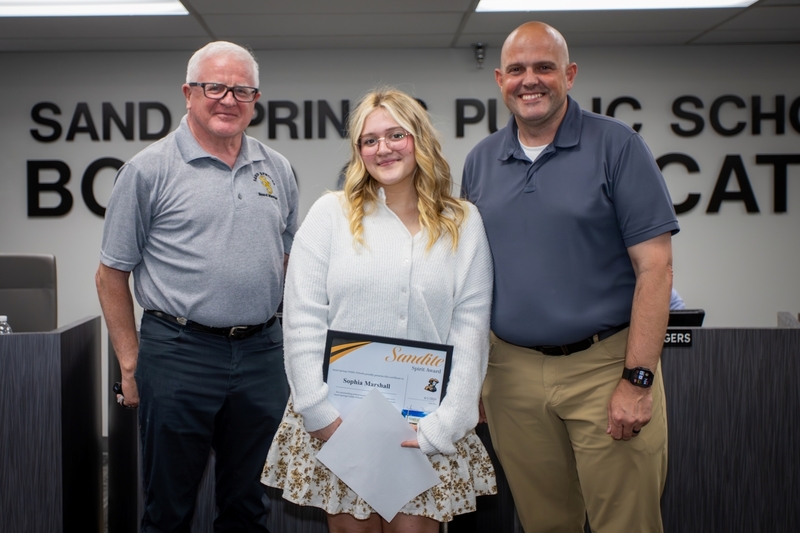 Student stands beside a board member and school administrator while holding a framed “Sandite Spirit Award” certificate during a Sand Springs Public Schools Board of Education recognition ceremony. All smile toward the camera in the board meeting room, with board members seated in the background and large wall lettering visible behind them.