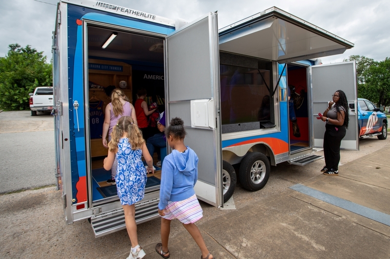 Children walk into the Oklahoma City Thunder Rolling Thunder Book Bus parked outside on a cloudy day. The mobile library has its doors open as students explore the Thunder-themed reading space inside, while an adult stands nearby supervising the visit.