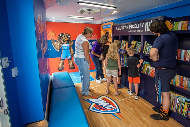 Children and adults browse books inside the Oklahoma City Thunder Rolling Thunder Book Bus. The mobile library features Oklahoma City Thunder branding, colorful bookshelves filled with children’s books, an OKC logo on the floor, and a mural of the Thunder mascot as students select books to read.