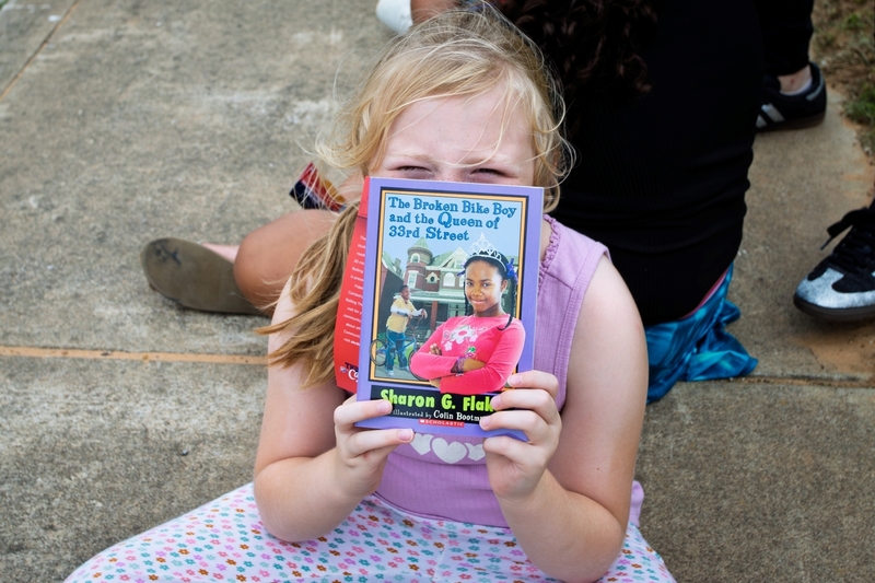 Young student sits outdoors holding a copy of “The Broken Bike Boy and the Queen of 33rd Street” by Sharon G. Flake in front of their face while smiling. The book cover is clearly visible, highlighting a moment of excitement and engagement with reading during a school literacy activity.
