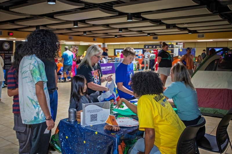 Families and students gather around activity tables at a school community event inside a cafeteria or commons area. Children participate in hands-on learning activities at a camping-themed booth decorated with a tent and campfire display, while informational tables and community organizations fill the background.