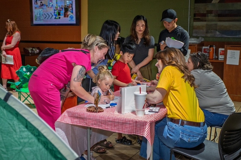 Families and students gather around an activity table at a literacy event while children complete hands on learning activities with guidance from volunteers. Parents and community members engage with educational resources and reading themed stations designed to encourage family participation and literacy development.