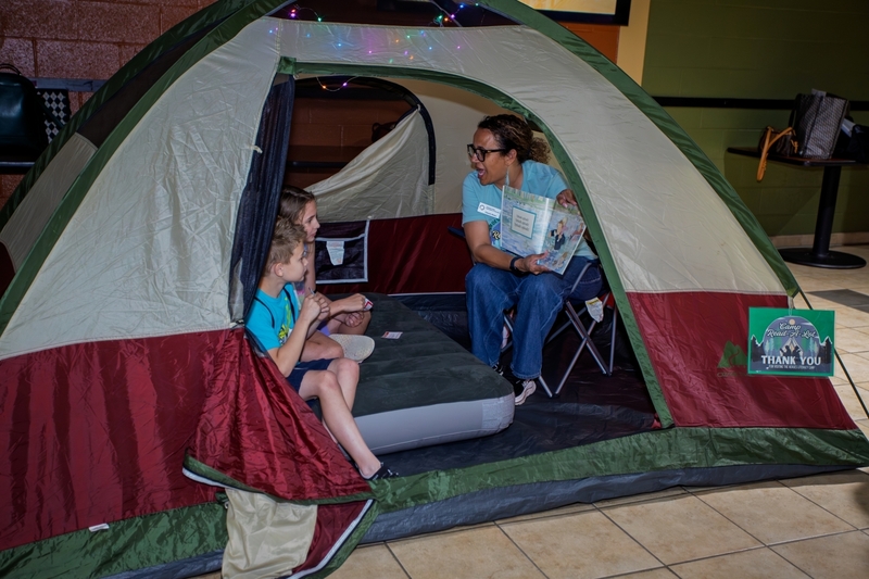 Educator reads aloud to two children seated inside a camping tent during a literacy themed family event. String lights, a storybook, and a cozy campsite setup create an inviting space that encourages reading, listening, and a love of books.